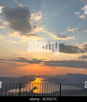 Atemberaubender Blick auf den Sonnenuntergang über die Küste, mit goldenem Sonnenlicht, das sich zwischen den Silhouetten der Berge unter einem dramatischen Himmel auf dem Meer spiegelt. Stockfoto