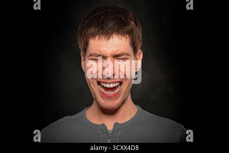 Ein junger Mann mit braunen Haaren, der ein graues Hemd trägt, schreiend oder lachend vor dunklem Hintergrund. Stockfoto