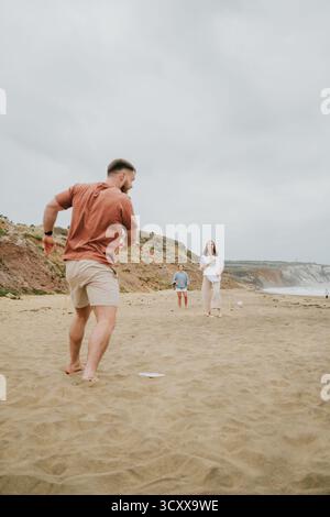Ein Mann in einem roten Hemd spielt Frisbee mit zwei anderen Menschen am Strand. Die Szene ist unbeschwert und unterhaltsam, während die Gruppe von Freunden ihre Zeit genießt Stockfoto