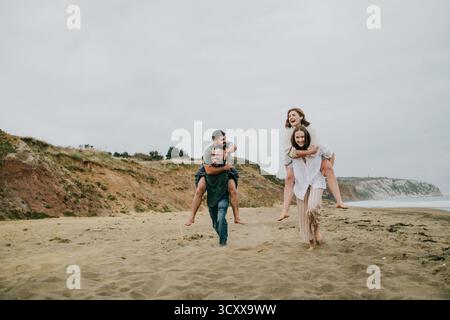 Eine Gruppe von Leuten spielt am Strand, mit einem Mann, der zwei Frauen auf dem Rücken trägt. Die Szene ist unbeschwert und lustig, während die Gruppe ihre genießt Stockfoto