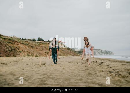 Eine Gruppe von Leuten spielt am Strand, wobei eine Frau von zwei Männern getragen wird. Die Szene ist unbeschwert und lustig, während die Gruppe ihre tim genießt Stockfoto