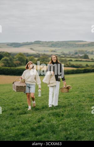 Zwei junge Frauen schlendern durch ein üppiges grünes Feld, von denen eine einen Korb trägt. Die Szene ist ruhig und ruhig, die Frauen genießen das B Stockfoto