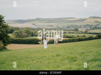 Zwei Frauen schlendern durch ein üppiges grünes Feld und genießen den wunderschönen Blick auf die sanften Hügel in der Ferne. Die Sonne scheint auf sie Stockfoto