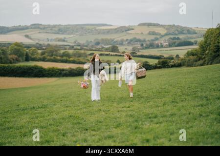 Zwei Frauen schlendern durch ein üppiges grünes Feld, umgeben von Blumen und einem Korb. Die Szene ist ruhig und ruhig, die Frauen genießen den Be Stockfoto