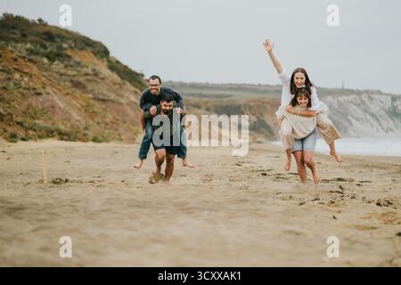 Eine vierköpfige Familie spielt am Strand eine Runde Tag. Der Mann trägt die Frau auf dem Rücken, während die anderen beiden rennen. Die Szene ist unbeschwert Stockfoto