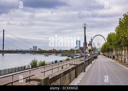 Von Bäumen gesäumter Rheinpromenadenweg mit entferntem Riesenrad und historischem Kirchturm unter bewölktem Himmel in Düsseldorf Deutschland Stockfoto