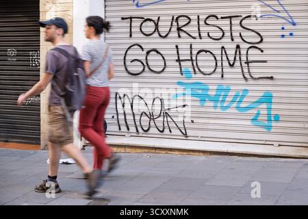 Ein junges Paar geht die Straße entlang, vorbei an einem Graffiti auf einem geschlossenen Ladenladen, auf dem steht „Touristen gehen nach Hause“ in Barcelona, Spanien Stockfoto