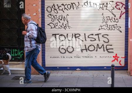 Ein Mann läuft die Straße entlang, vorbei an einem Graffiti auf einem geschlossenen Ladenladen, auf dem steht „Touristen gehen nach Hause“ in Barcelona, Spanien Stockfoto