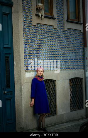 Eine Frau in Fliedermantel und Baskenmütze steht an einer mit Azulejos bedeckten Wand in Porto, Portugal, während ein Hund vom Fenster hinunter blickt. Die Szene f Stockfoto