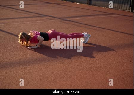 Starke Frau in rosa Leggings und Top, die Liegestütze auf einem Outdoor-Sportplatz bei Sonnenuntergang macht. Stockfoto
