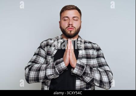 Ein junger Mann mit Bart meditiert ruhig mit den Händen zusammen. Er trägt ein kariertes Hemd und ist in einem ruhigen Innenraum, mit geschlossenen Augen, Fokus Stockfoto