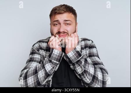 Ein junger Mann mit einem verspielten Ausdruck ballt seine Fäuste nahe am Kinn und zeigt Gefühle von Aufregung oder Nervosität. Er steht in einem einfachen in Stockfoto