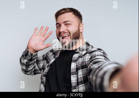 Ein fröhlicher Mann, der ein kariertes Hemd trägt, winkt begeistert, während er ein Selfie macht. Das Ambiente ist lässig und spiegelt ein entspanntes Zuhause wider Stockfoto