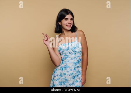 Eine junge Frau mit dunklem Haar steht selbstbewusst und trägt ein hellblaues Blumenkleid. Sie lächelt und zeigt spielerisch, vor einem schlichten beigen Hintergrund Stockfoto
