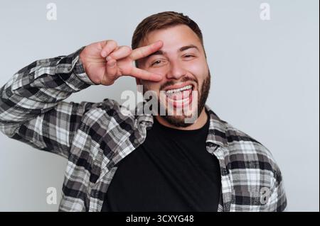 Ein Mann lächelt breit, während er mit der Hand ein Friedenszeichen macht. Er trägt ein kariertes Hemd über einem schwarzen T-Shirt und ist in einem hellen, neutralen Setting. Sein Stockfoto