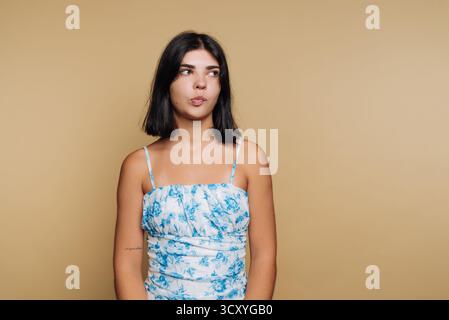 Eine junge Frau steht in einem Studio und trägt ein Blumenoberteil. Sie schaut mit einem nachdenklichen Ausdruck auf ihrem Gesicht weg. Der neutrale Hintergrund fügt den Fokus hinzu Stockfoto