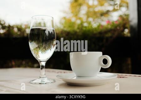Weiße Porzellantasse mit heißem Tee oder starkem türkischen schwarzen Kaffee oder Americano und einem Glas Wasser, serviert auf einem Café-Tisch im Freien bei Tageslicht. Stockfoto