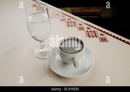 Weiße Porzellantasse mit starkem türkischen schwarzen Kaffee oder Americano und einem Glas Wasser, serviert auf einem Café-Tisch im Freien bei Tageslicht. Stockfoto