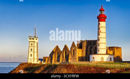 Der hohe Leuchtturm Phare de Saint-Mathieu steht neben den Überresten einer alten Steinabtei. Die untergehende Sonne wirft warmes Licht und hebt die Küste hervor Stockfoto