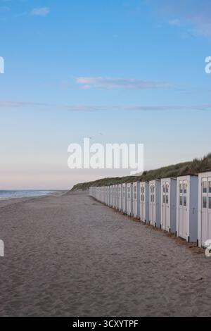 Eine lange Reihe von weißen Strandhütten, die sich in die Ferne entlang des riesigen Sandstrandes erstrecken und von grasbewachsenen Dünen unter einem klaren Abendhimmel begleitet werden Stockfoto