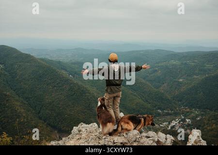 Eine glückliche Frau hebt ihre Arme auf dem Gipfel des Mount Kablar, Serbien, mit zwei Hunden australischen und deutschen Schäferhunden neben ihr, die die Freiheit des Mannes genießen Stockfoto