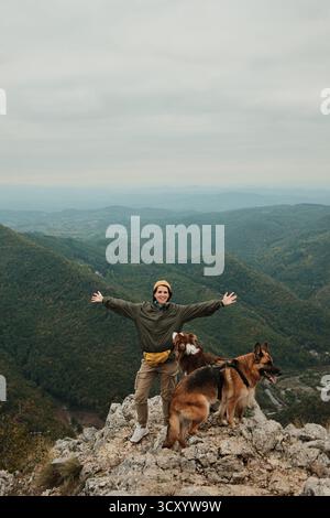 Eine glückliche Frau hebt ihre Arme auf dem Gipfel des Berges Kablar, Serbien, mit ihren beiden Hunden Australian und German Shepherd neben ihr, und genießt die Freiheit Stockfoto