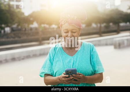 Glückliche afrikanische Seniorin mit Smartphone im Freien mit Stadt im Hintergrund, die traditionelle Kleidung trägt - fröhlicher älterer Lebensstil und Technologie Stockfoto