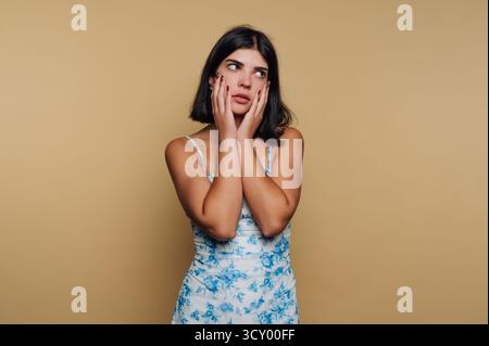 Eine junge Frau mit kurzen Haaren posiert vor beigefarbenem Hintergrund in einem hellblauen Blumenkleid. Sie hat einen nachdenklichen Blick und zeigt ihre Hand auf ihrem fa Stockfoto