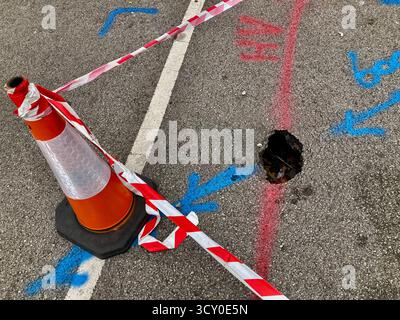 Ein Beispiel eines Schlaglochs auf einer Straße, abgesperrt mit einem Verkehrskegel und rot-weißem Warnband Stockfoto