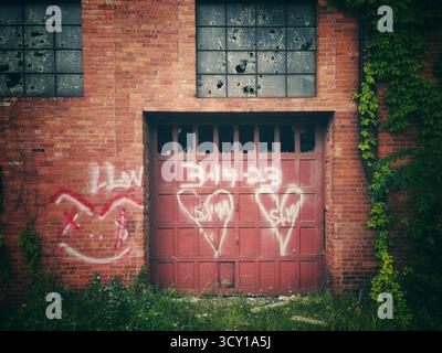 Altes industrielles Backsteingebäude mit rotem Garagentor, bedeckt mit Graffiti und umgeben von bewachsenen Pflanzen und Weinstöcken Stockfoto