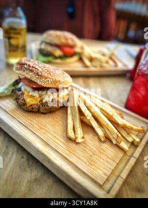 Zwei Burger mit Pommes Frites auf Holzbrettern auf einem Tisch sowie erfrischende Getränke in zwangloser Atmosphäre Stockfoto