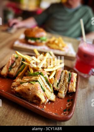 Nahaufnahme eines köstlichen Club-Sandwiches mit knusprigen Pommes, serviert auf einem Teller, mit einem Burger und Getränk im Hintergrund auf einem Holztisch Stockfoto