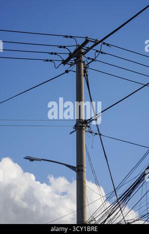 Ein Versorgungsmast mit mehreren Stromleitungen und einer Straßenlaterne vor einem blauen Himmel mit Wolken Stockfoto