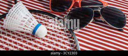 Ein Tennisschläger, ein Badminton-Shuttlecock und eine Sonnenbrille stehen auf einem rot-weiß gestreiften Tisch Stockfoto