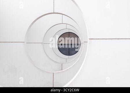 Ein markantes minimalistisches architektonisches Detail mit einem weißen konzentrischen Spiraldesign, das zu einem runden Fenster mit einem dunklen metallischen Gitter führt. Stockfoto