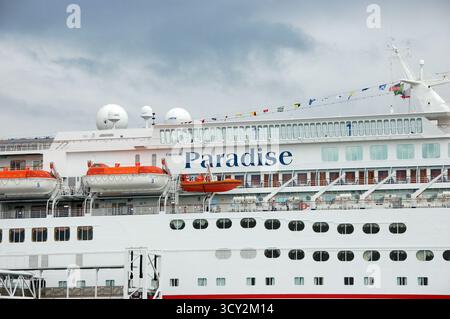 Das Kreuzfahrtschiff Carnival Paradise legte am Hafen unter bewölktem Himmel an, bereit für die Abfahrt mit farbenfrohen Flaggen und ruhigem Meereswasser Stockfoto
