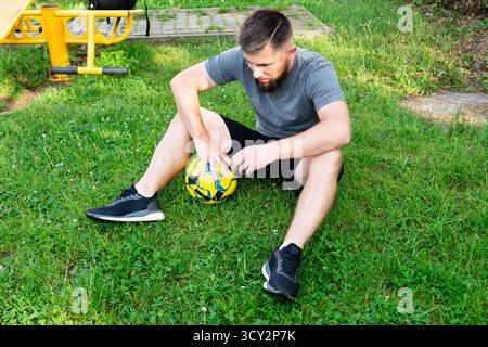 Ein bärtiger Mann sitzt auf dem Gras und hält einen gelb-blauen Fußball. Er scheint nach einem Spiel oder Training eine Pause einzulegen. Ein Fitnessraum Stockfoto