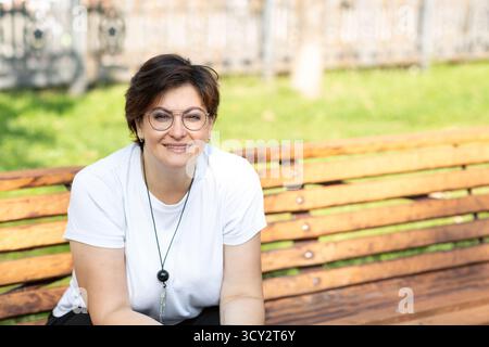 Eine schöne Frau mittleren Alters mit Brille und kurzen dunklen Haaren sitzt auf einer Bank Stockfoto