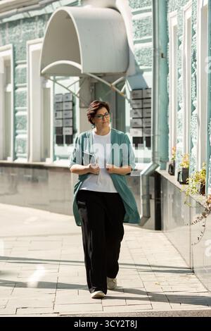 Eine Frau mittleren Alters mit Brille geht die Straße hinunter mit einem Notizbuch in der Hand Stockfoto
