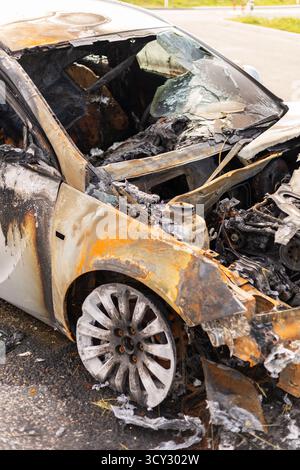 Verbrannte Wrackteile weisen erhebliche Schäden durch verkohltes Metall und Asche auf. Das Fahrzeug steht in einem Industriegebiet neben der Straße und weist Schilder auf Stockfoto