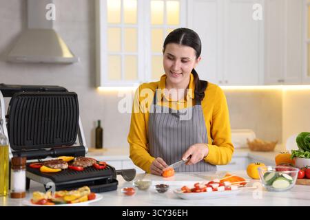 Frau, die Pfeffer schneidet, während sie Fleisch und Gemüse auf einem elektrischen Grill in der Küche kocht Stockfoto