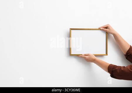 Frau, die einen leeren Diplomrahmen an der weißen Wand hängt, Großaufnahme. Modell für Design Stockfoto