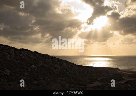Goldenes Licht breitet sich über den Himmel und das Meer aus, während die Sonne über den Küstenklippen untergeht. Wellen schlagen sanft gegen die Küste und schaffen eine ruhige Szene in der Th Stockfoto