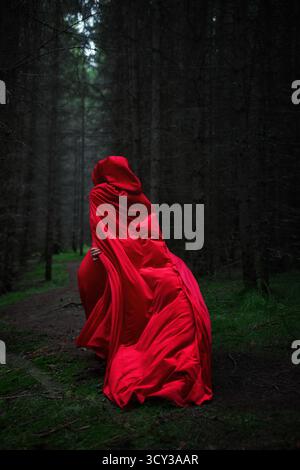 Man sieht die Frau, die durch einen dichten, schwach beleuchteten Wald geht, in einen langen roten Mantel gehüllt. Die Atmosphäre fühlt sich mystisch an, während sich der Nebel zwischen den Bäumen niederlässt, cre Stockfoto