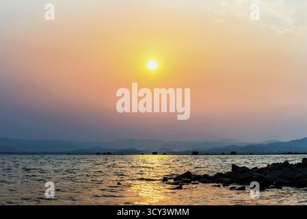 Schöne Naturlandschaft gelbe Sonne in der orange Himmel See spiegelt das goldene Sonnenlicht auf der Wasseroberfläche bei Sonnenuntergang, Fischfarm Käfige Berg b Stockfoto