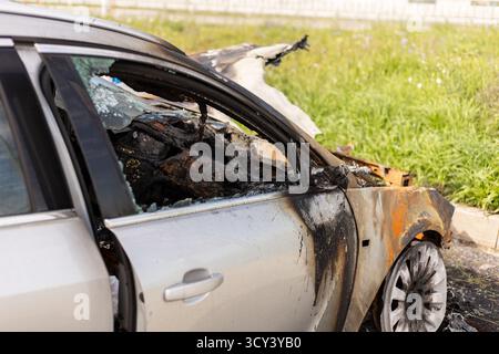 Am Straßenrand ist ein schwer durch Feuer beschädigtes Fahrzeug zu sehen. Das Metall ist verbrannt und geschwärzt, zwischen grünem Gras und Resten des verbrannten Fahrzeugs Stockfoto