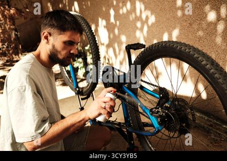 Konzentrierter Mann, der sein blaues Mountainbike pflegt, indem er draußen Schmiermittel auf die Kette spritzt. Konzept von Pflege, Pflege und aktivem Leben. Stockfoto
