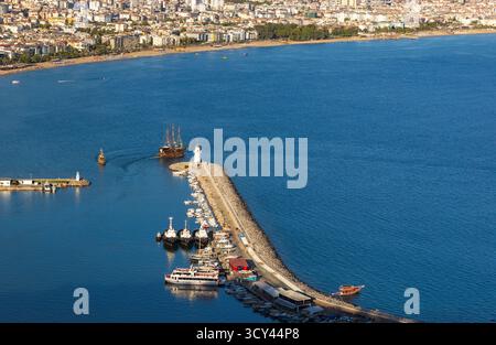 Hafenszene von Alanya mit einem Steinbruch, einem weißen Leuchtturm, vertäuten Booten und einer fernen Stadtküste. Klares, blaues Wasser und sonniges Wetter sorgen dafür Stockfoto