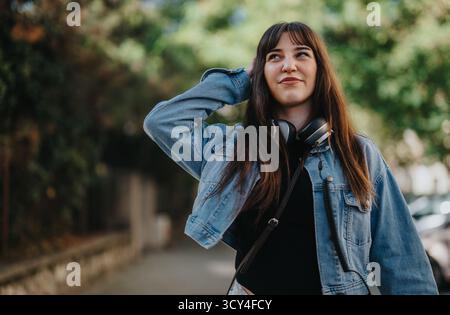 Lächelnde junge Frau, die draußen eine Jeansjacke mit Kopfhörern um den Hals trägt Stockfoto