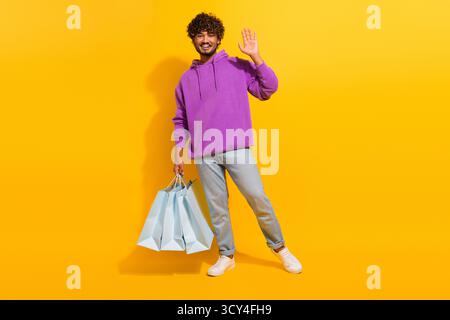 Junger Mann in lässigem lila Hoodie, der Einkaufstaschen hält und die Hand vor hellgelbem Hintergrund winkt Stockfoto